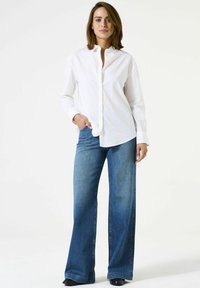 Vrouw die met één hand in haar zak staat, gekleed in een wit knoopshirt en een blauwe wijde jeans, tegen een plain witte achtergrond.