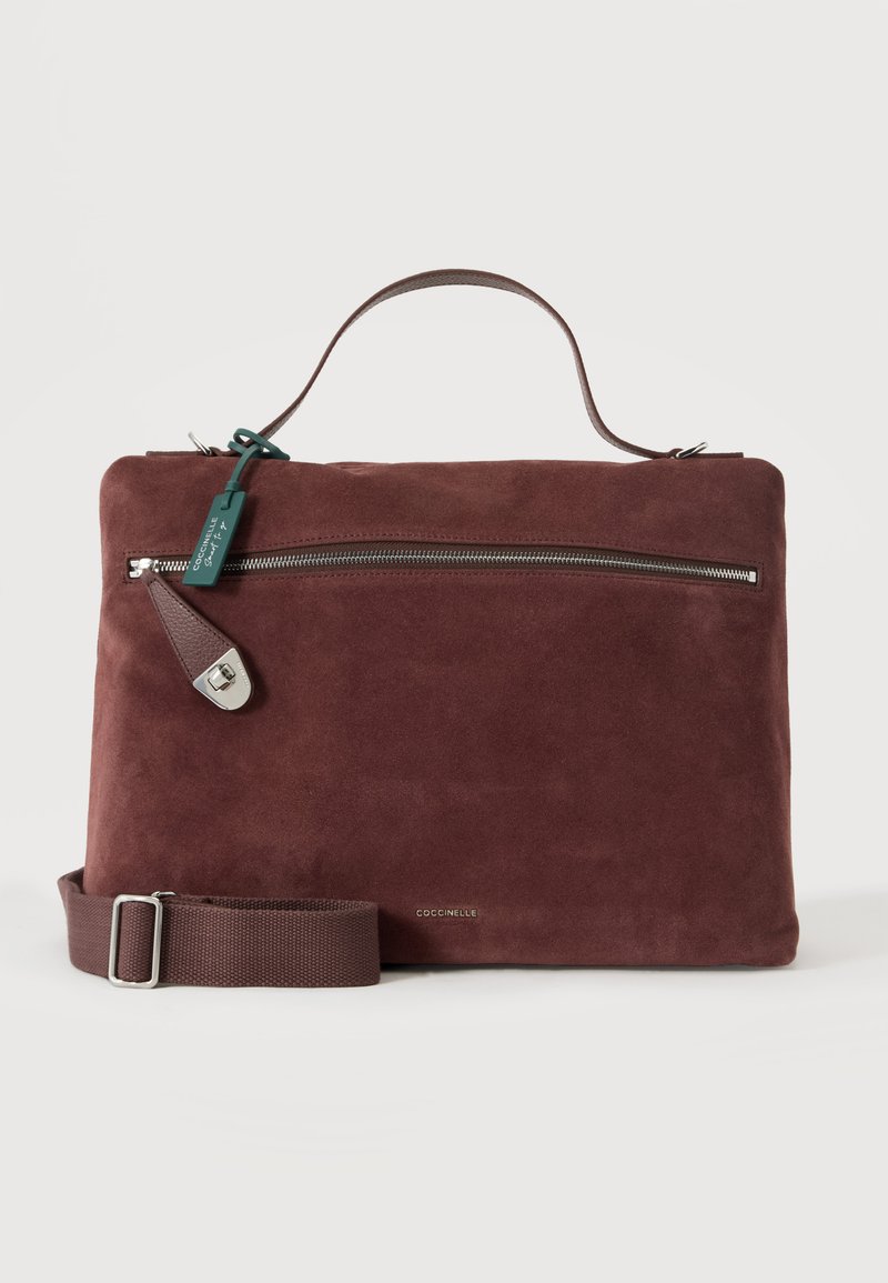 Mallette en daim marron foncé avec poignée supérieure, poche avant zippée, bandoulière réglable et logo de marque en métal sur fond uni.