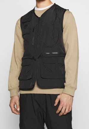 Gilet en tissu noir avec plusieurs poches, fermeture éclair à l'avant et design léger. Porté par-dessus une chemise à manches longues beige.