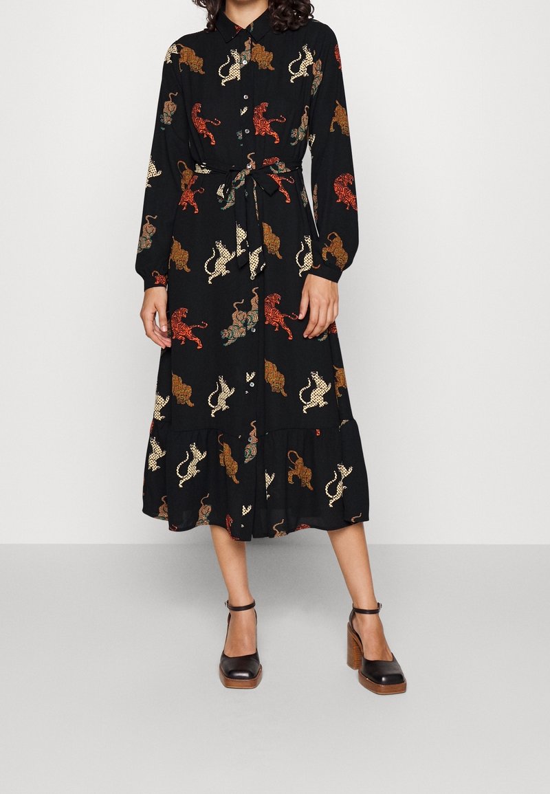 Femme portant une robe midi noire à manches longues avec des imprimés tigres colorés, nouée à la taille, associée à des talons noirs à bride à la cheville.