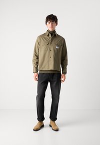 Olive green shirt jacket with two chest pockets, black buttons, and a tag. Worn over a dark turtleneck and black jeans, with rolled cuffs.