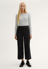 Kvinna med långt blont hår iklädd en vit långärmad randig topp, svarta vida cropped-byxor och svarta snörskor, ståendes.