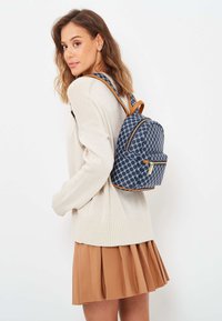 Marinblå ryggsäck med beige detaljer, dragkedjestängning och en framficka. Bärs på ryggen, ihop med en krämfärgad tröja och en brun veckad kjol.