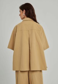 Beige oversized skjorte med korte ærmer, detalje med rygfold og en struktureret krave. Lavet af glat stof, der falder til midt-hofte længde.