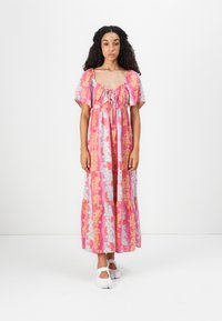 Kvinde med langt krøllet hår iført en pink blomster-maksikjole med puffærmer og hvide sko, stående foran en ensfarvet hvid baggrund.