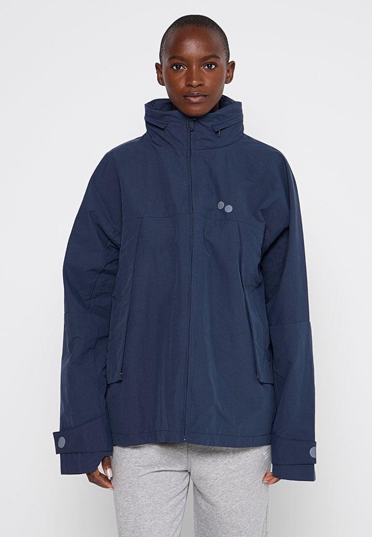 Marineblaue wasserdichte Jacke mit hohem Kragen, Reißverschluss und zwei Fronttaschen. Hat weite Ärmel und runde Akzentdetails.