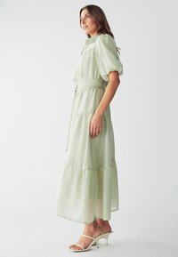 Vestido maxi verde claro hecho de una tela transparente, con mangas abullonadas, un cinturón atado en la cintura y un diseño de falda en capas.