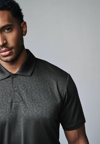 Sort polo-shirt med et tekstureret geometrisk mønster, kortærmet og med en klassisk krave; har tre knapper foran.