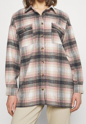 Femme portant une chemise boutonnée à manches longues à motif à carreaux rose, noir et blanc et un pantalon beige.