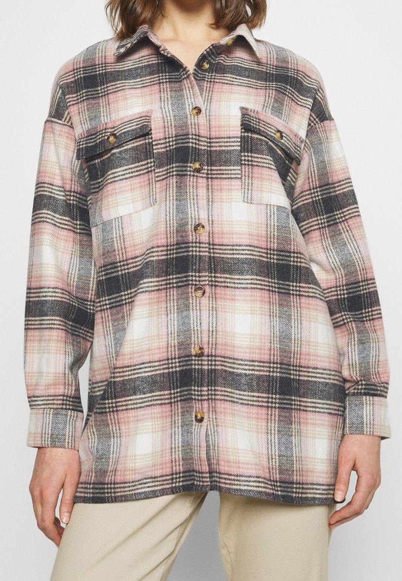 Femme portant une chemise boutonnée à manches longues à motif à carreaux rose, noir et blanc et un pantalon beige.