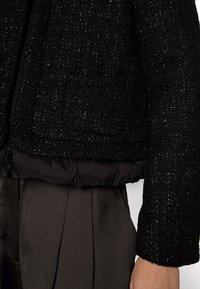 Black textured jacket with a quilted black lining, featuring a structured design, slightly cropped length, and long sleeves.