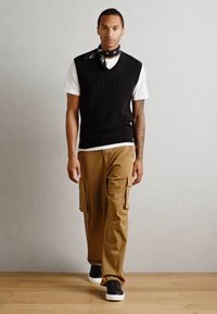 Vesta neagră cu dungi, purtată peste un tricou alb, asortată cu pantaloni cargo bej cu buzunare laterale și adidași negri cu talpă albă.