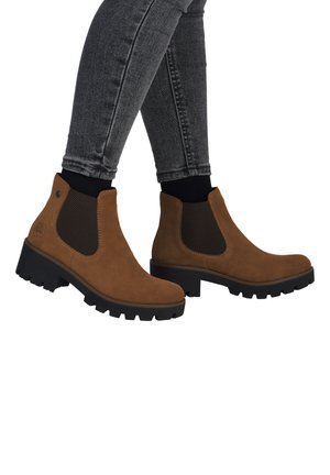 Persoon draagt bruine suède Chelsea-laarzen met zwarte elastische zijpanelen en dikke zwarte zolen, gecombineerd met zwarte sokken en donkergrijze skinny jeans.