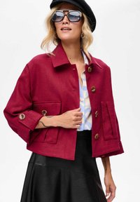 Rode linnen jack met een kort ontwerp, grote voorzakken en knoopsluitingen. Gecombineerd met een blauw patroon top en een zwarte rok.