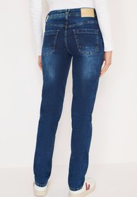 Jean bleu foncé à coupe slim, avec une taille haute, deux poches arrière et une texture douce. Le logo de la marque est visible sur la ceinture.