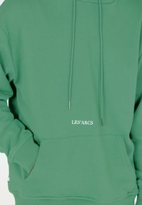 Grüne Kapuzenjacke mit einer Fronttasche und Kordeln. Mit aufgedrucktem Text "LES' ARCS" in Weiß. Hergestellt aus weichem, fleeceähnlichem Material, lockere Passform.