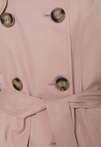 Rosa trenchcoat i tyg med mörka runda knappar och ett knutet midjeband, med detaljerad sömnad och knapphål.