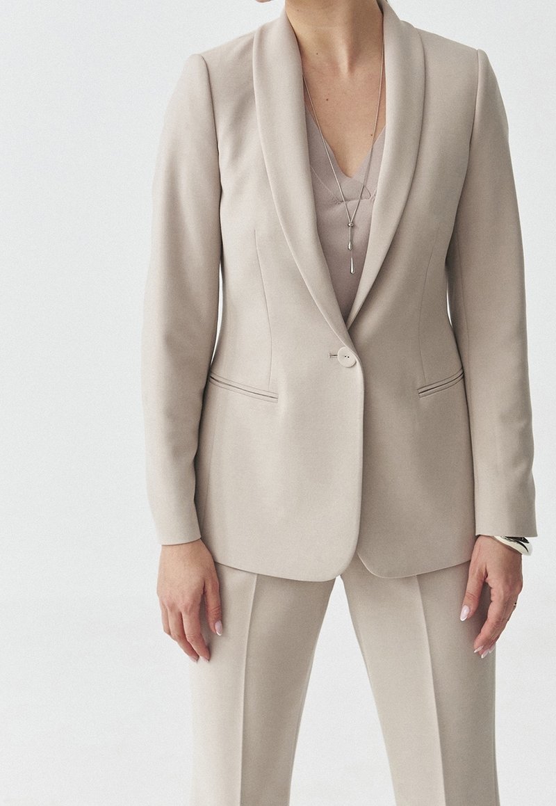 Costume beige avec une veste ajustée et un pantalon. La veste présente un revers aigu, une fermeture à un bouton et des poches latérales discrètes.