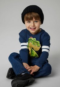 Maglietta a maniche lunghe di colore blu navy con grafica di dinosauro, strisce bianche sulle maniche, abbinata a jeans blu e scarpe nere, e un berretto a maglia nero.