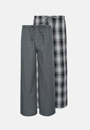 Homme debout pieds nus portant un t-shirt gris à manches longues et un pantalon à carreaux noir et blanc avec cordon, sur fond uni.