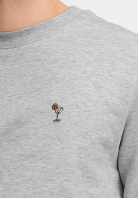 Sudadera gris hecha de una tela suave, con un pequeño diseño bordado de un vaso de cóctel con una rodaja de naranja.