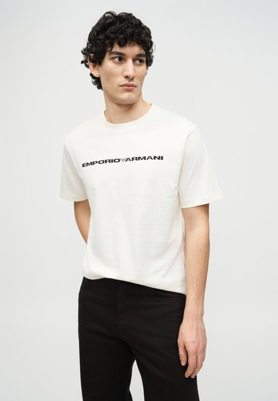 Νέος άνδρας με σκούρα σγουρά μαλλιά, φορώντας λευκό μπλουζάκι Emporio Armani και μαύρο παντελόνι, κοιτάζει προς τα αριστερά του σε απλό φόντο.