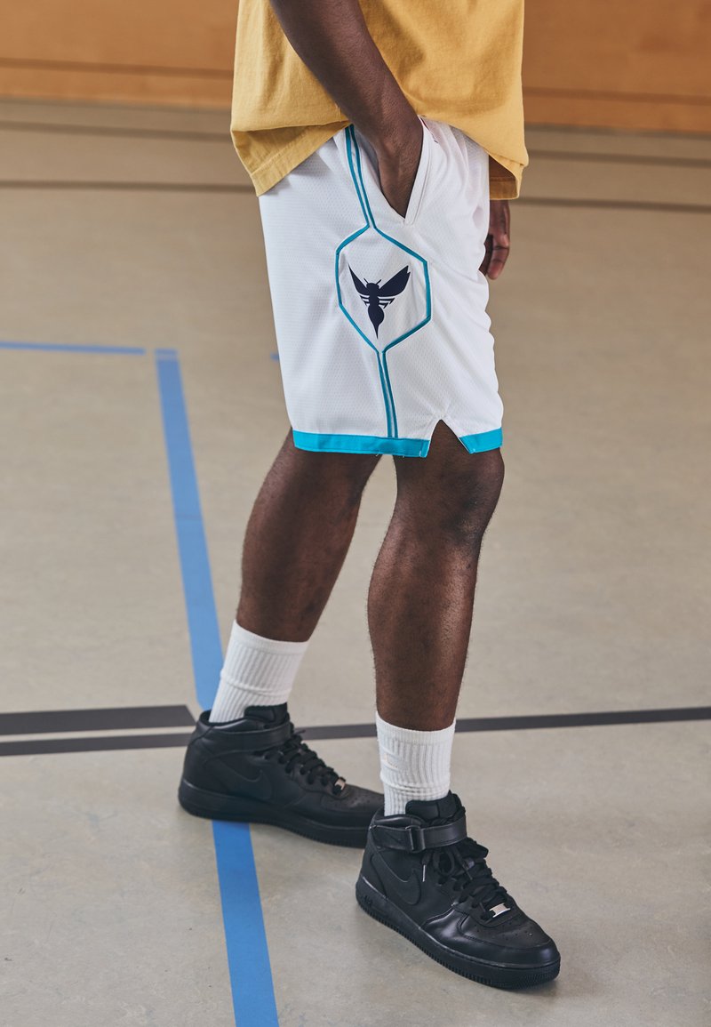 White athletic shorts with blue accents, featuring a black logo, mesh texture, and side pockets, paired with black high-top sneakers.