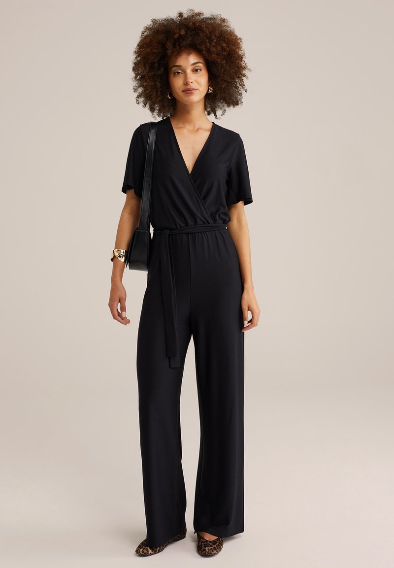 Zwarte jumpsuit met een wikkelontwerp, korte mouwen en een ceintuur in de taille. Gladde stof; ruime pasvorm, aangevuld met flats in luipaardprint.