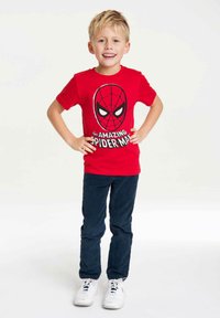 Red cotton t-shirt featuring a Spider-Man graphic on the front, paired with dark blue pants and white sneakers. Child stands confidently.