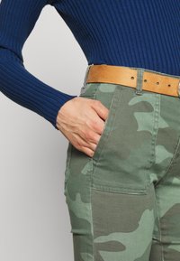 Mano descansando en el bolsillo delantero de un pantalón de camuflaje verde con cinturón marrón, combinado con una camiseta de manga larga azul con canalé.
