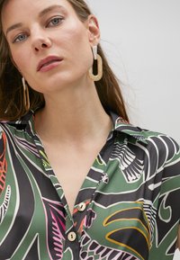 Green patterned blouse with floral and abstract designs, featuring a collar and three visible buttons. Gold statement earrings complete the look.