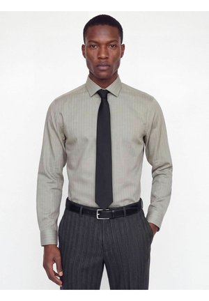Homme portant une chemise gris clair à manches longues avec des rayures discrètes, une cravate noire, une ceinture noire et un pantalon gris foncé à fines rayures, debout sur un fond blanc.