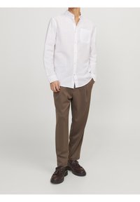 Camisa de lino blanca de mangas largas con un bolsillo en el pecho, combinada con pantalones marrones ajustados y calzado marrón. Detalles de botones simples visibles.