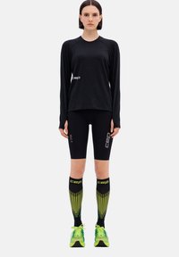Maglietta sportiva nera a maniche lunghe, pantaloni corti neri e calze a compressione nere e gialle fino al ginocchio. Scarpe da corsa verde brillante con dettagli riflettenti.