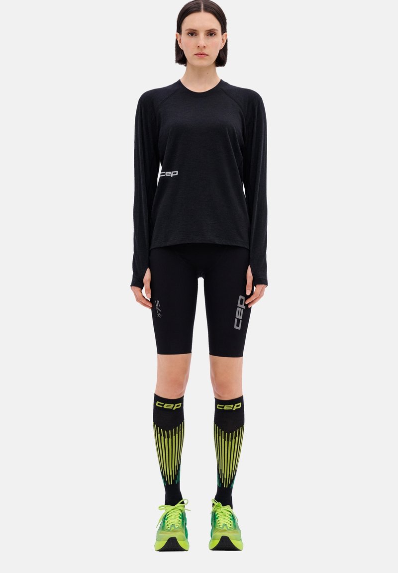 Maglietta sportiva nera a maniche lunghe, pantaloni corti neri e calze a compressione nere e gialle fino al ginocchio. Scarpe da corsa verde brillante con dettagli riflettenti.