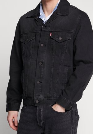 Mand iført sort denimjakke med knapper, hånd i lommen og en delvist synlig blå og hvid ternet skjorte under.