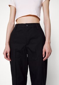 Femme portant un pantalon noir taille haute avec fermeture par boutons et un top court à manches courtes rose clair, debout devant un fond blanc.