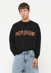 Felpa nera con una grande grafica arancione "REPLAYING". Presenta una vestibilità comoda e polsini a costine. Abbinata a jeans in denim azzurro chiaro.