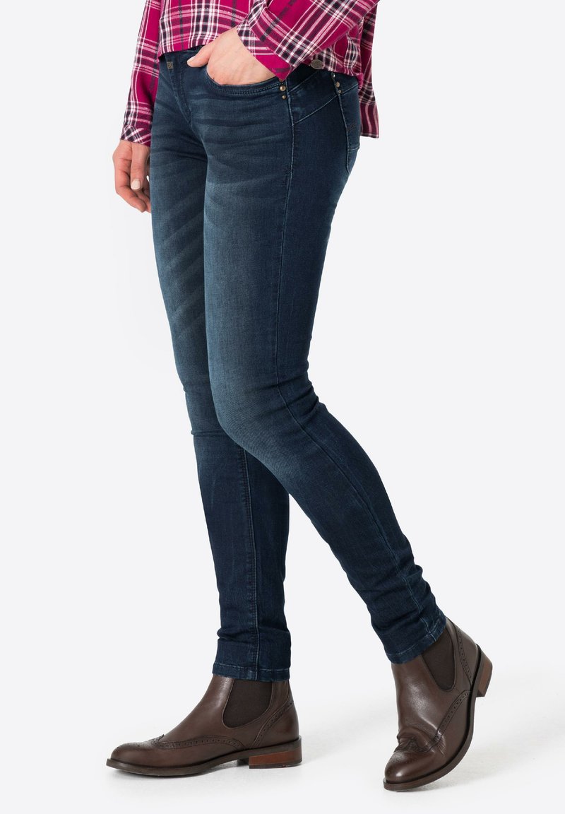 Dark blue skinny jeans with a smooth texture, highlight stitching, and pocket accents, paired with brown leather ankle boots.