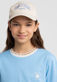 Jasnoniebieska koszula z długim rękawem z białym kołnierzykiem i kwiatowym logo; beżowa czapka z haftem "BIG STAR JEANS" w niebieskim kolorze; naszyjnik z pereł.