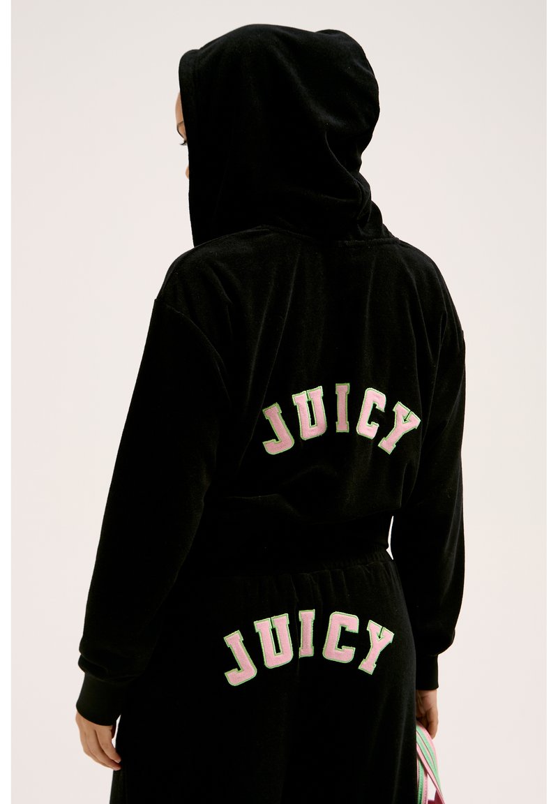 Set di felpa e pantaloni tuta in velluto nero con la scritta "JUICY" in lettere ricamate rosa e verdi sul retro. Include un cappuccio e una vita elastica.
