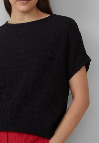 Haut noir à manches courtes avec un motif texturé et ondulé. Col rond, coupe décontractée et détails de couture visibles. Assorti avec un tissu rouge en dessous.