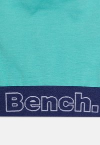Turkis stoff med en glatt tekstur, med et mørk marineblått midjebånd som har "Bench." brodert i hvitt.