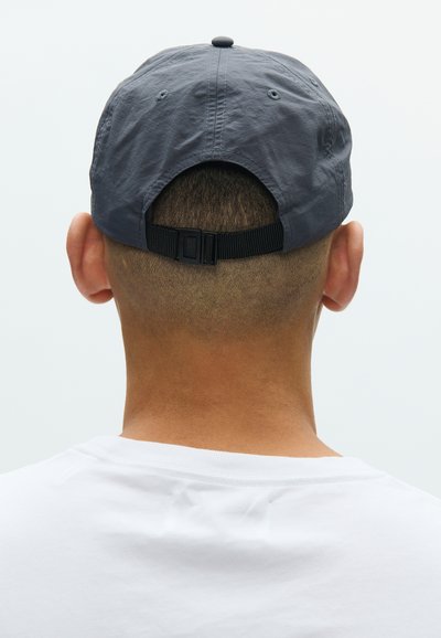 Γκρι καπέλο με στρογγυλό στέμμα, ρυθμιζόμενο λουρί στο πίσω μέρος, υφή υφάσματος με ελαφριά λάμψη, φορεμένο πάνω από λευκό πουκάμισο.