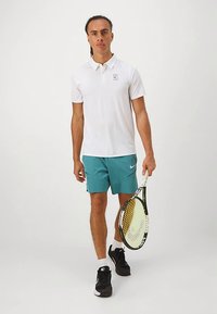 Biele polo tričko s malým logom, kombinované s tyrkysovými šortkami a čiernymi teniskami, držiace tenisovú raketu. Hladká látka a športový strih.