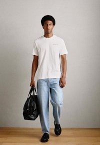 Weißes T-Shirt mit dezentem Text, hellblaue Jeans und schwarze Leder-Dufflebag. Das Modell trägt schwarze Schuhe und steht auf einem Holzboden vor einer grauen Wand.
