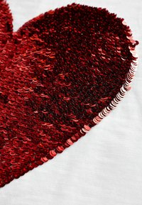 Röd paljetthjärta på en vit tygbakgrund, med glänsande cirkulära paljetter som är tätt arrangerade, vilket skapar en texturerad yta.
