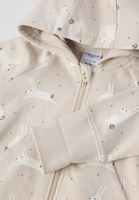 Beige baby hoodie i blødt stof med et mønster af kaniner og blomster. Har en fuld lynlås, hætte og ribbede manchetter. Pyntet med lysebrune prikker.