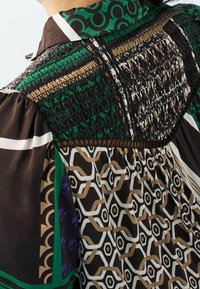 Prenda con cuello y mangas abullonadas que presenta patrones geométricos y fruncidos en colores verde, marrón, negro, blanco y beige sobre la tela.