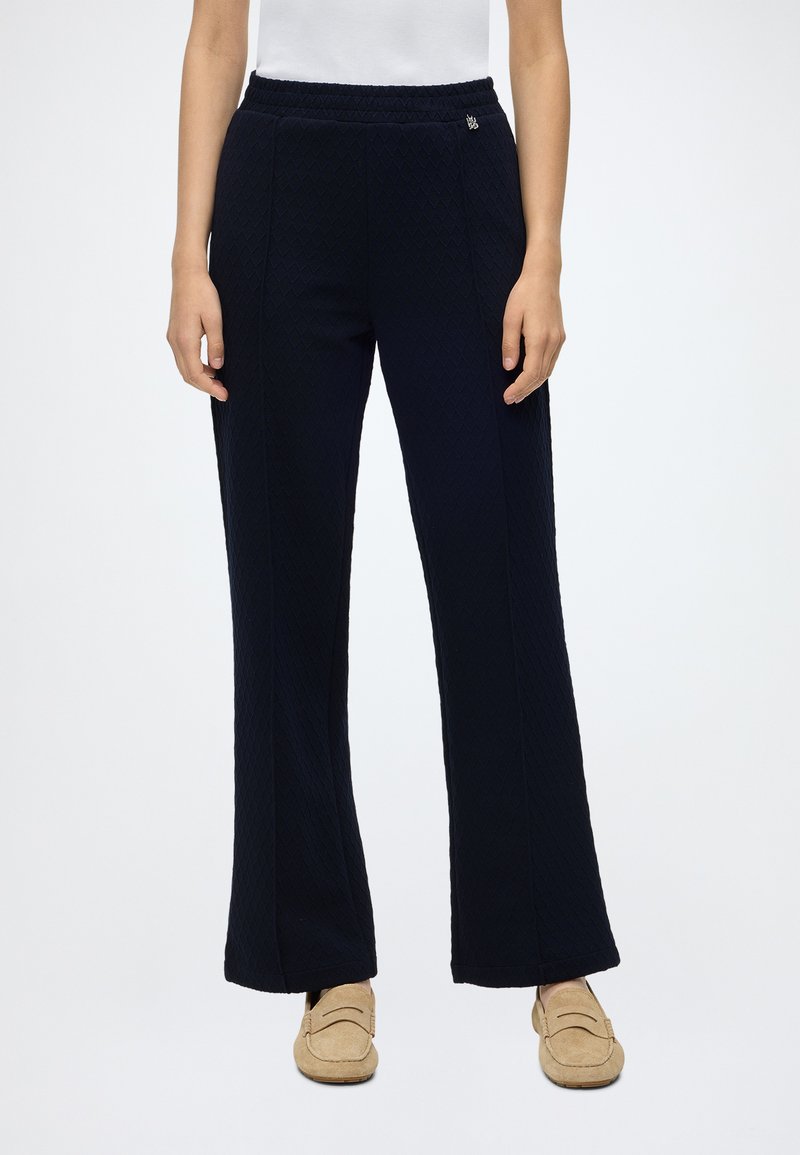 Femme portant un pantalon noir texturé à jambes larges et des mocassins beiges, debout sur un fond blanc uni.